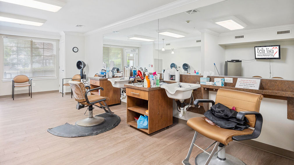 A bright, modern salon with styling chairs, mirrors, hair washing sinks, and shelves holding hair products and tools. Large windows let in natural light, and a TV is mounted on the wall displaying "Bella Vita.