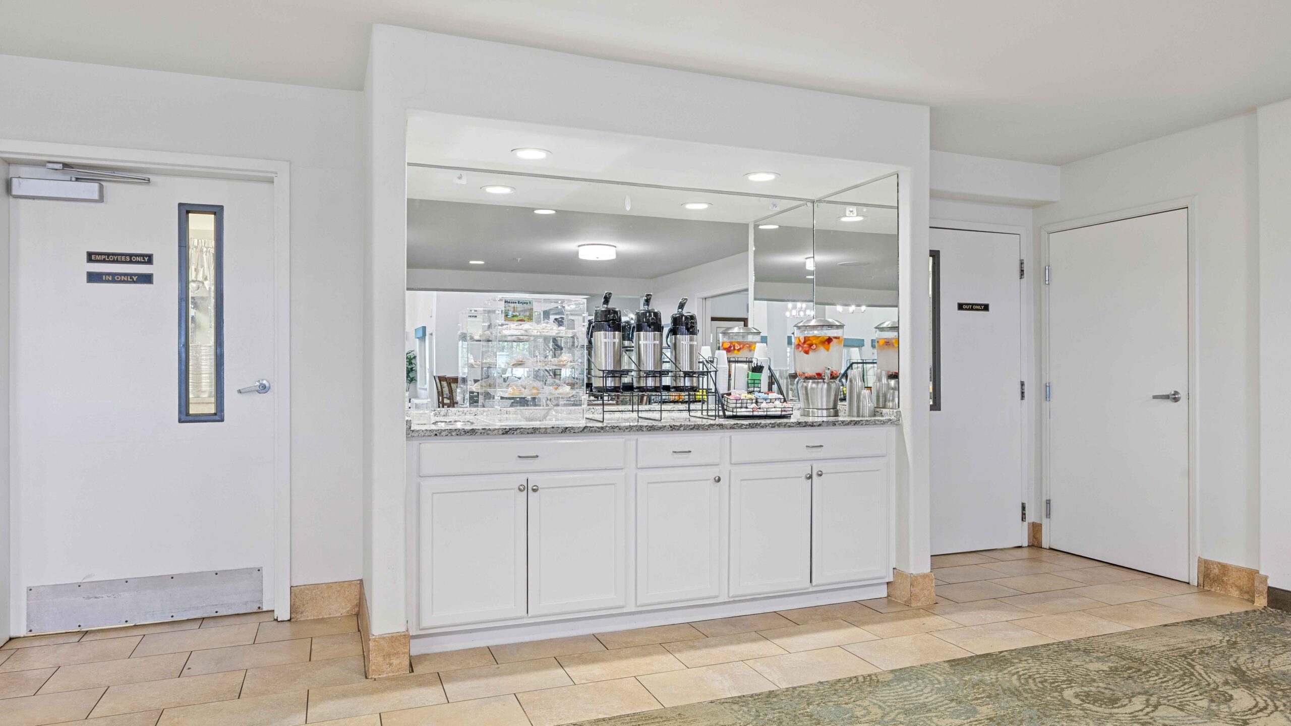 Breakfast buffet area with white cabinets, a mirrored backsplash, and various breakfast items such as juice dispensers and coffee urns arranged on the countertop. There are two doors on either side of the buffet.