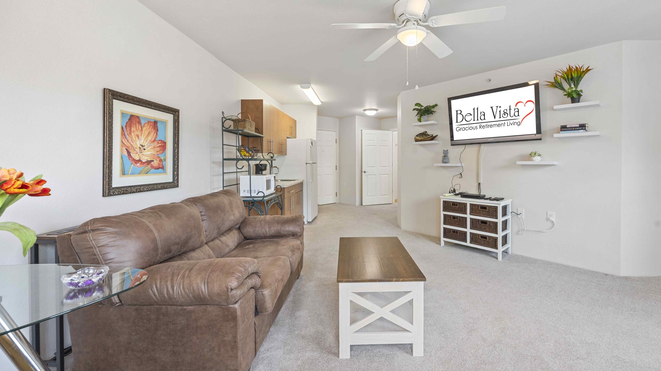 A cozy living area with a brown couch, wooden coffee table, wall art, and plants. A wall-mounted TV displays "Bella Vista." The room has beige carpet, white walls, and a ceiling fan. Kitchen area is visible in the background.