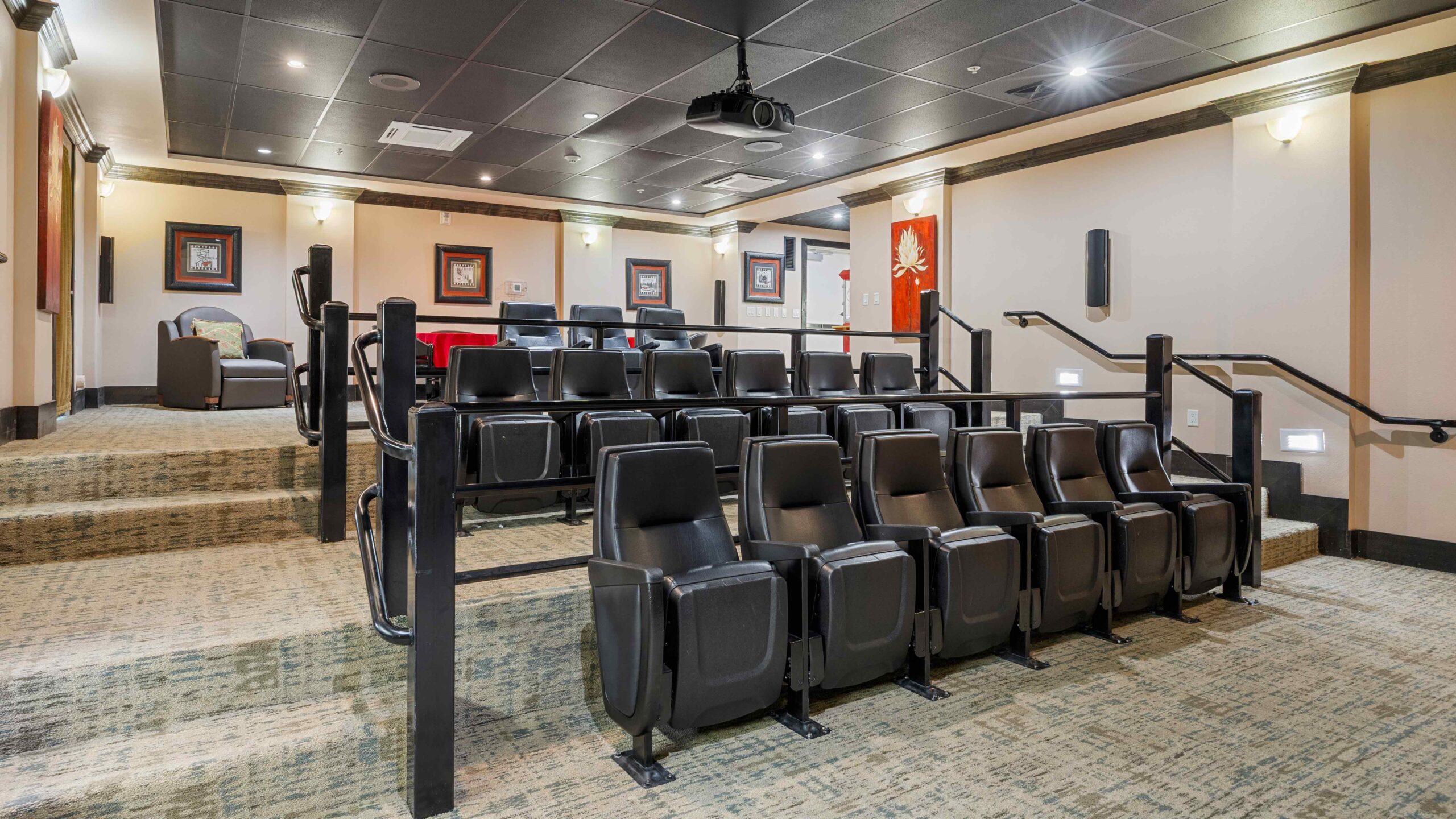 A home theater room with three rows of black reclining seats, a projector on the ceiling, framed pictures on the beige walls, and soft lighting along the aisles.