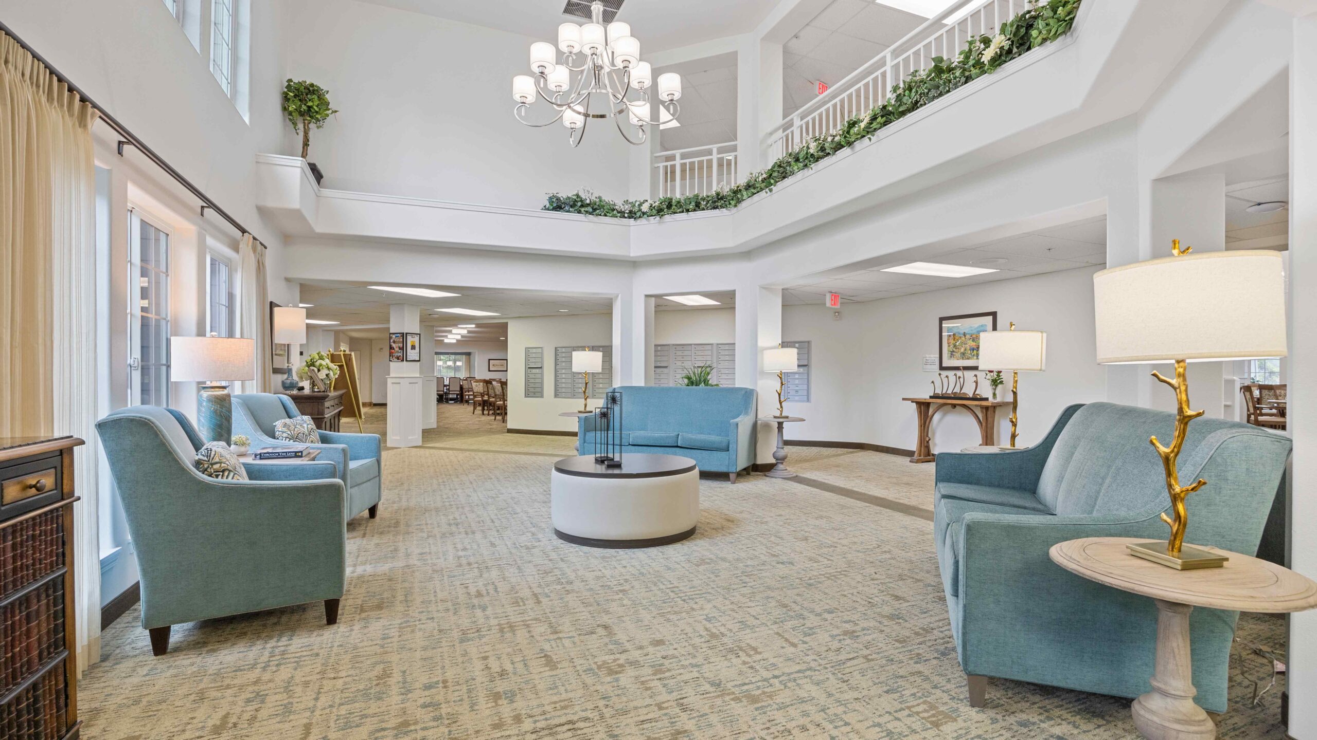 A bright, spacious lounge with blue armchairs, a round ottoman, large windows, and indoor plants. A chandelier hangs from the high ceiling, and a second-floor balcony overlooks the area.