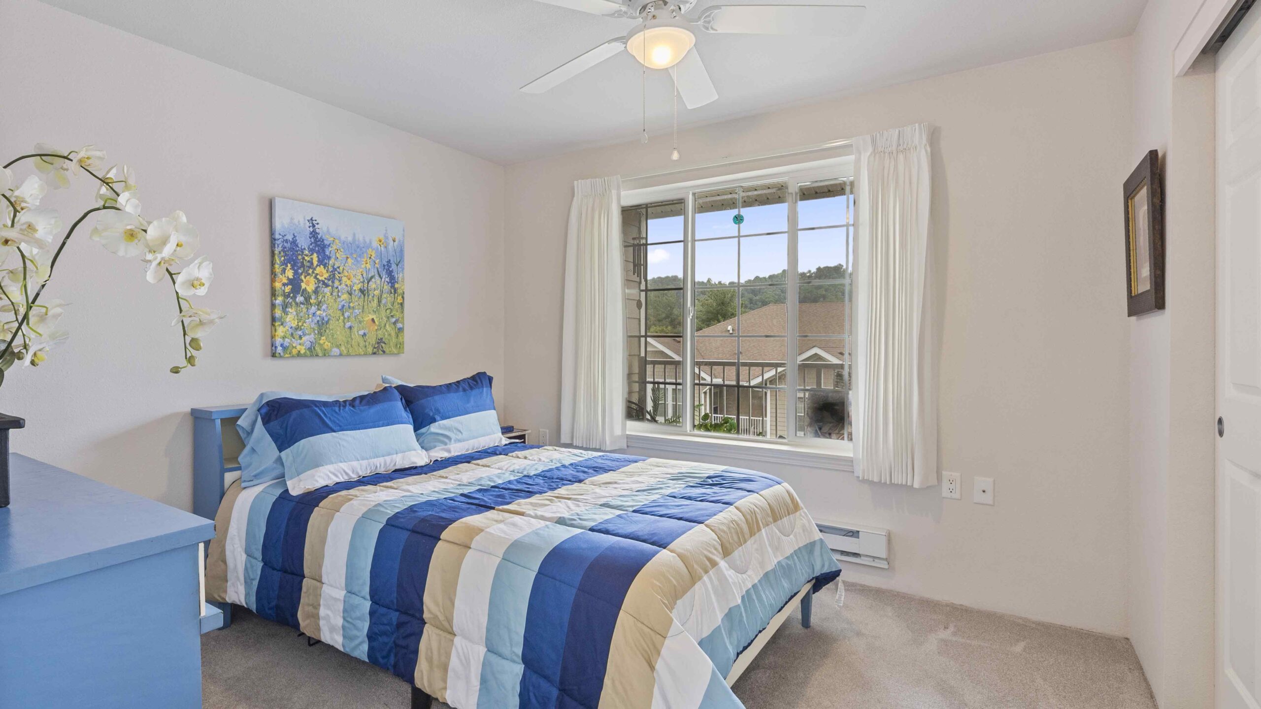 A bright bedroom with a blue striped bedspread, blue nightstand, floral wall art, and a large window with white curtains overlooking a suburban neighborhood. A ceiling fan is above the bed.