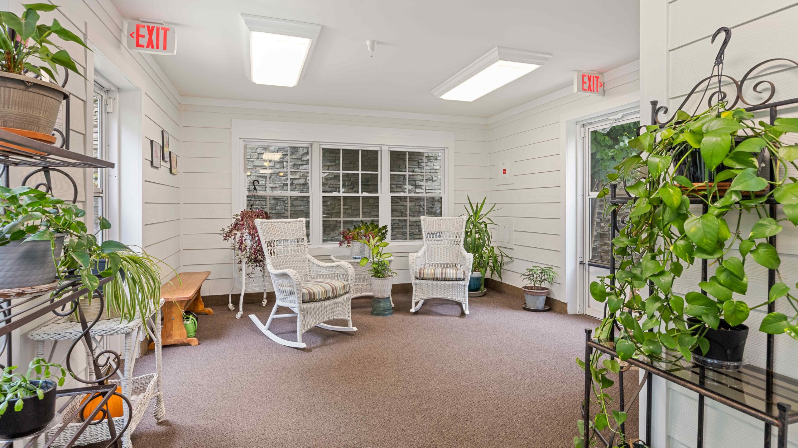 A bright sunroom with white walls, two white wicker rocking chairs with cushions, potted plants on shelves and the floor, a wooden bench, a large window, and exit signs above two doors.