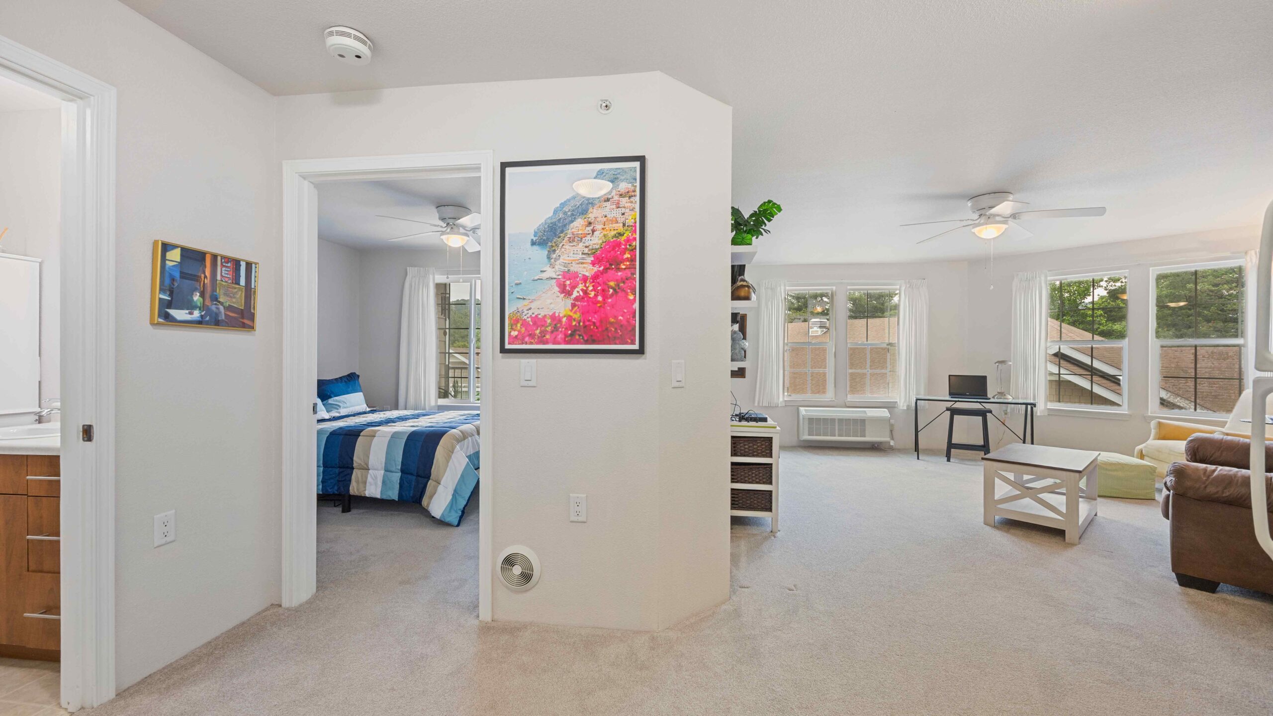 A bright apartment interior with a bedroom to the left, a bathroom partially visible, and a spacious living room with large windows, ceiling fans, and modern furniture. A colorful framed picture hangs on the wall.
