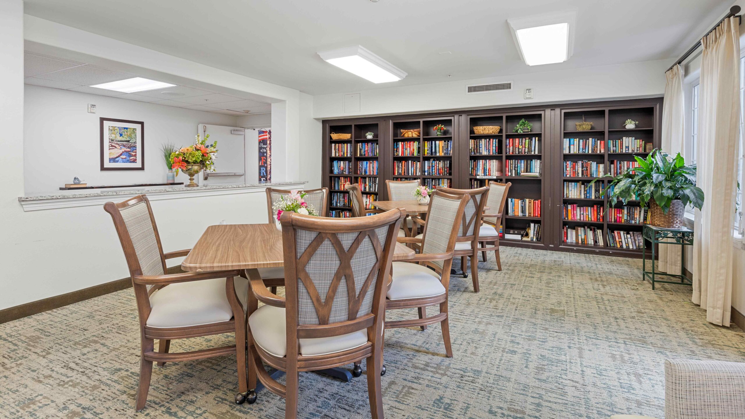 A bright, cozy library room with wooden bookshelves filled with books, round tables and chairs, a large potted plant, and natural light coming in through curtained windows.