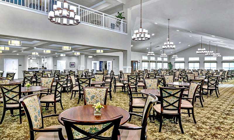 A spacious dining area with multiple round tables and chairs arranged neatly. The room features high ceilings, large windows, chandeliers providing ample light, and a patterned carpeted floor. Some tables have small flower centerpieces.