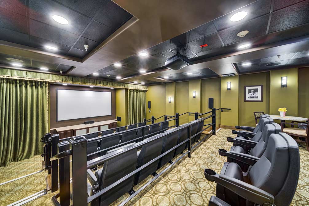 A private home theater with plush individual seating and a large screen showing an old western movie. The room is decorated with green curtains, soft lighting, and framed pictures on the wall. The carpet has a detailed pattern, and the seating is arranged in rows.