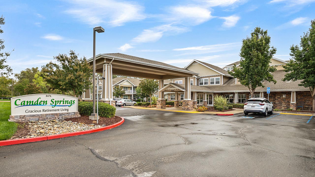 A retirement living facility with a covered driveway entrance, a sign reading "Camden Springs Gracious Retirement Living," landscaping, parked cars, and a bright, partly cloudy sky.