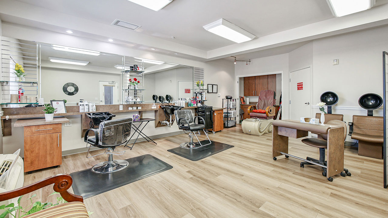 A modern, well-lit salon with styling stations, chairs, mirrors, hair dryers, a manicure table, and a pedicure chair. The space has light wood floors and neutral-colored walls, creating a clean and welcoming environment.