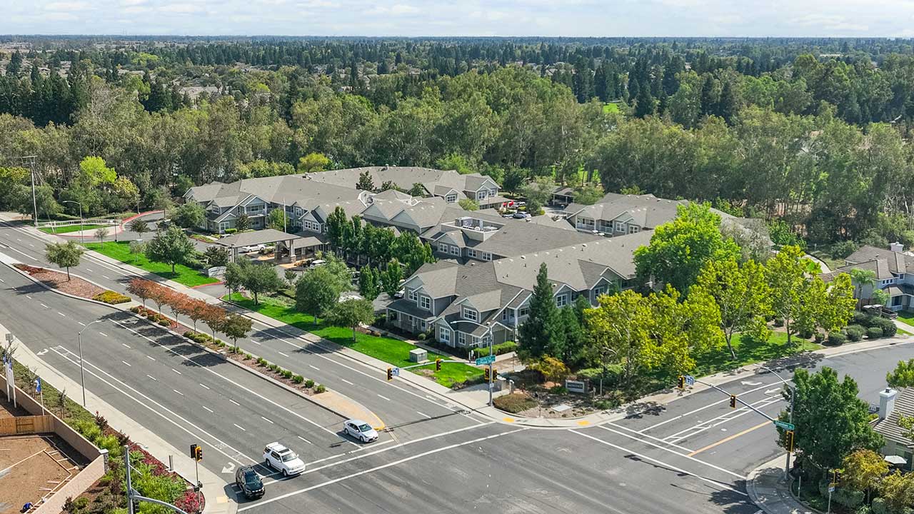 Aerial view of a suburban neighborhood with tree-lined streets, several gray-roofed apartment buildings, and a large intersection with cars and traffic lights in the foreground. Lush greenery surrounds the area.