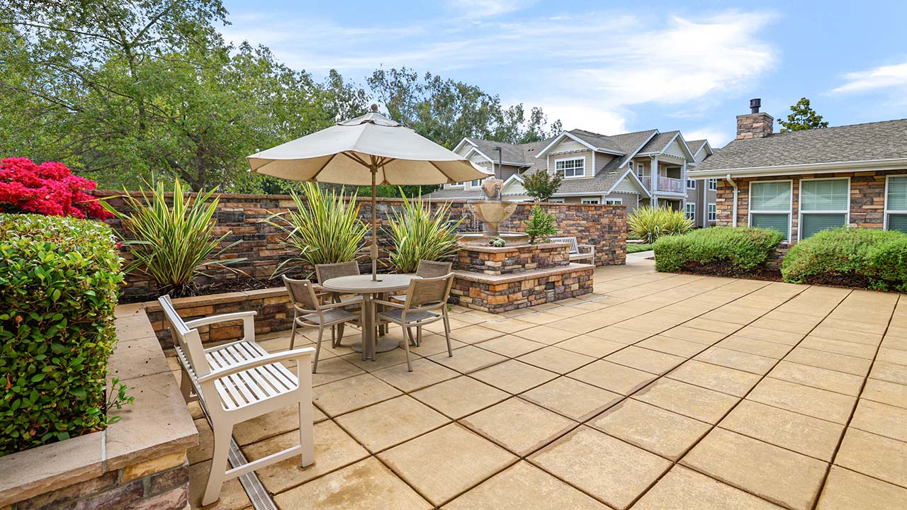 A spacious, tiled patio with a table, chairs, and an umbrella sits next to a stone wall with plants and flowers. Benches and bushes line the area, with houses and trees in the background under a blue sky.