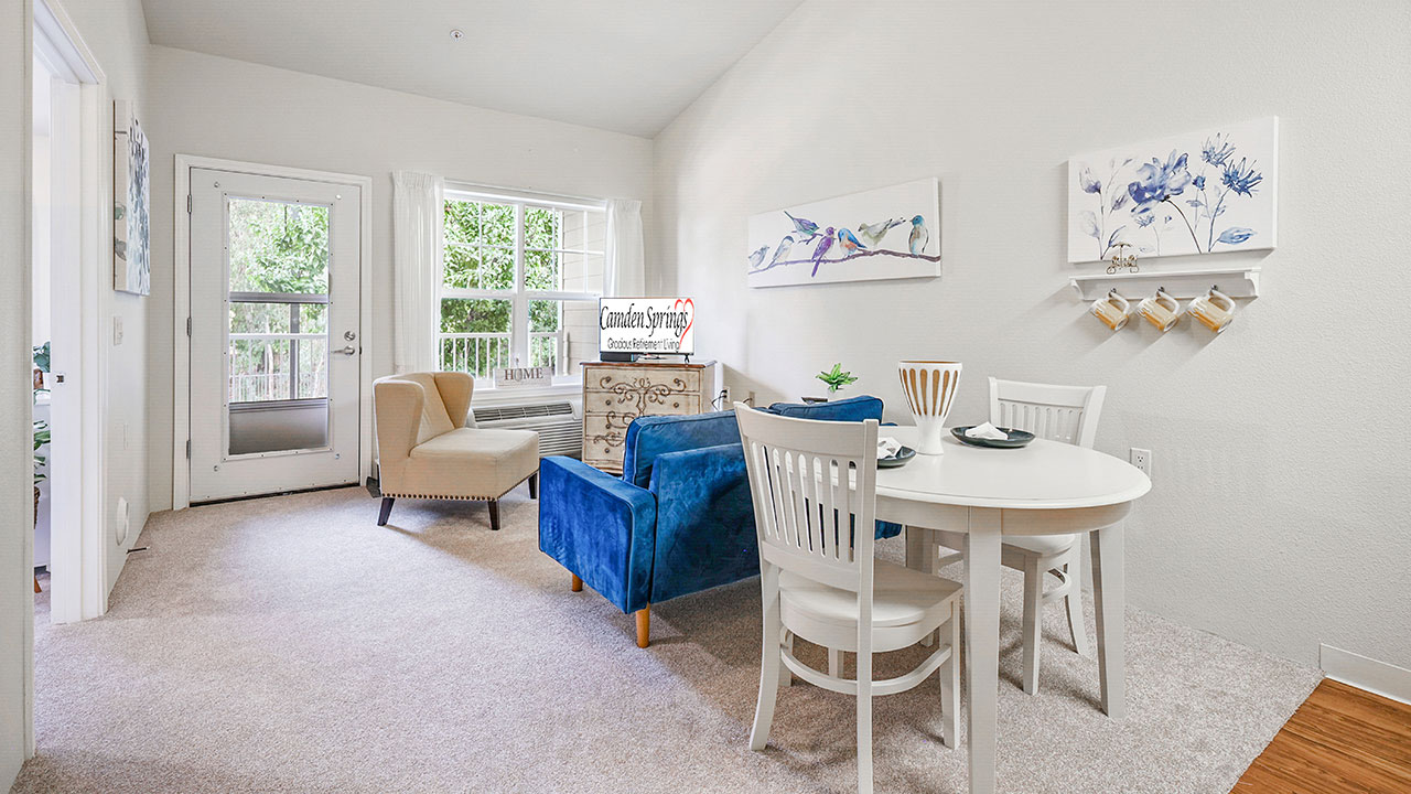 A cozy, bright living and dining area with a round white table, four chairs, a blue armchair, a beige chair, and wall art. Large windows and a glass door let in natural light and offer views of greenery outside.