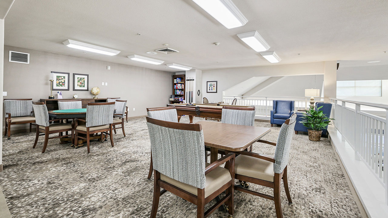 A spacious, well-lit game room with tables and chairs, a bookshelf, framed artwork, and large windows; the room features neutral tones, carpet flooring, and a railing along one side.