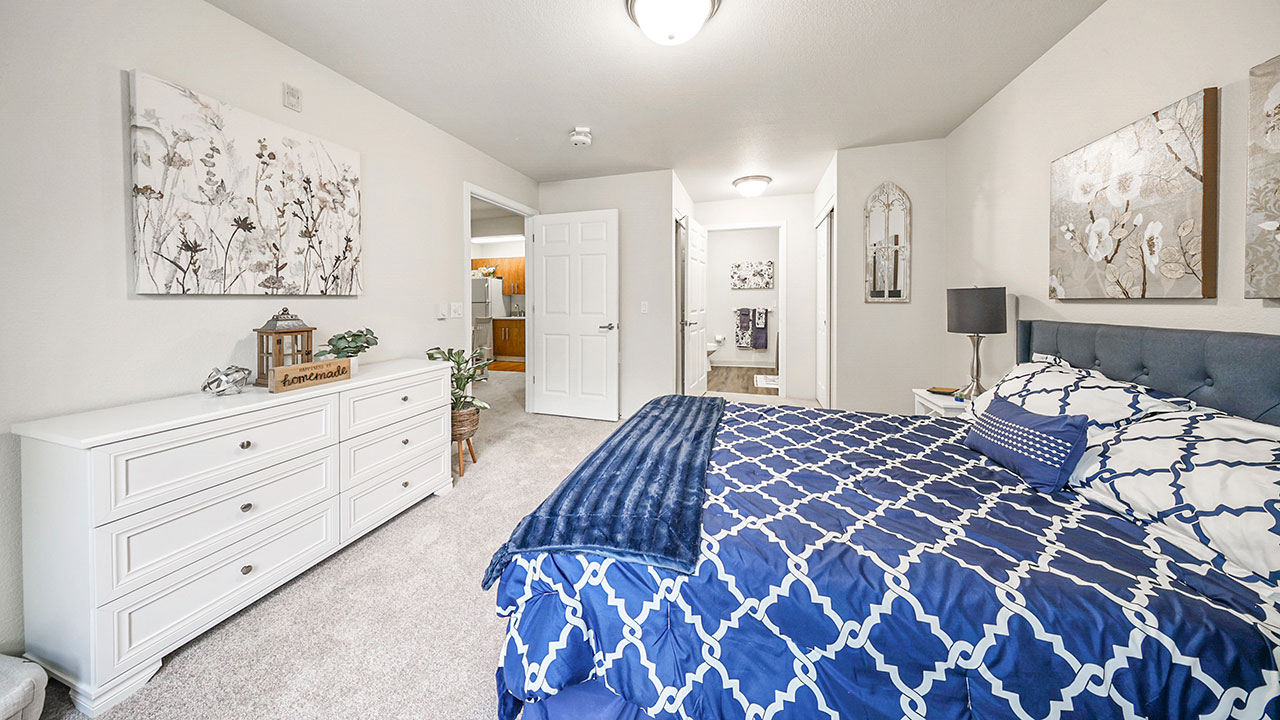 A bedroom with a blue and white patterned bedspread, white dresser, wall art, and neutral walls. An open door reveals a hallway leading to a bathroom and another room in the background.
