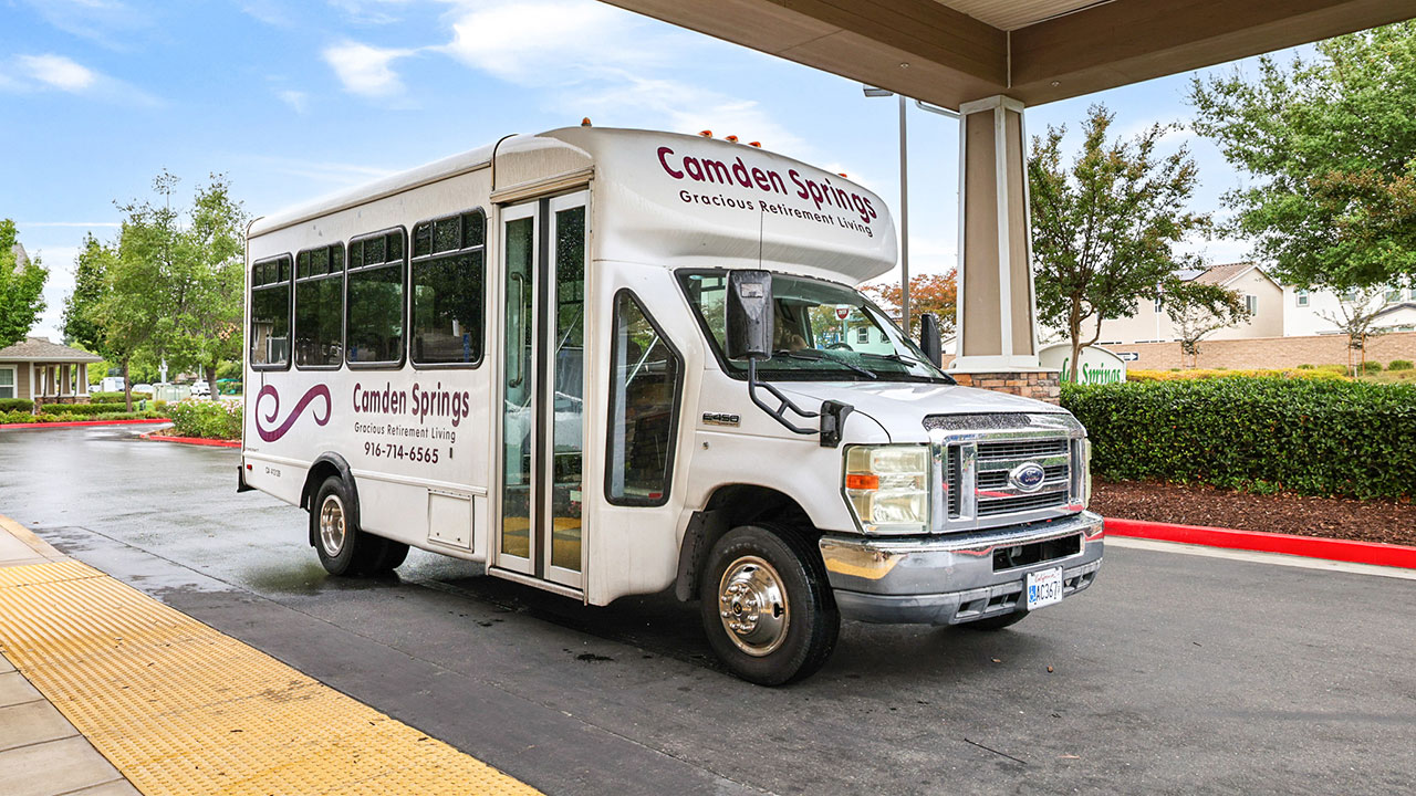 A white Camden Springs shuttle bus is parked at a curb under a canopy, with trees and shrubs in the background. The bus has large windows and signage displaying the Camden Springs name and contact information.