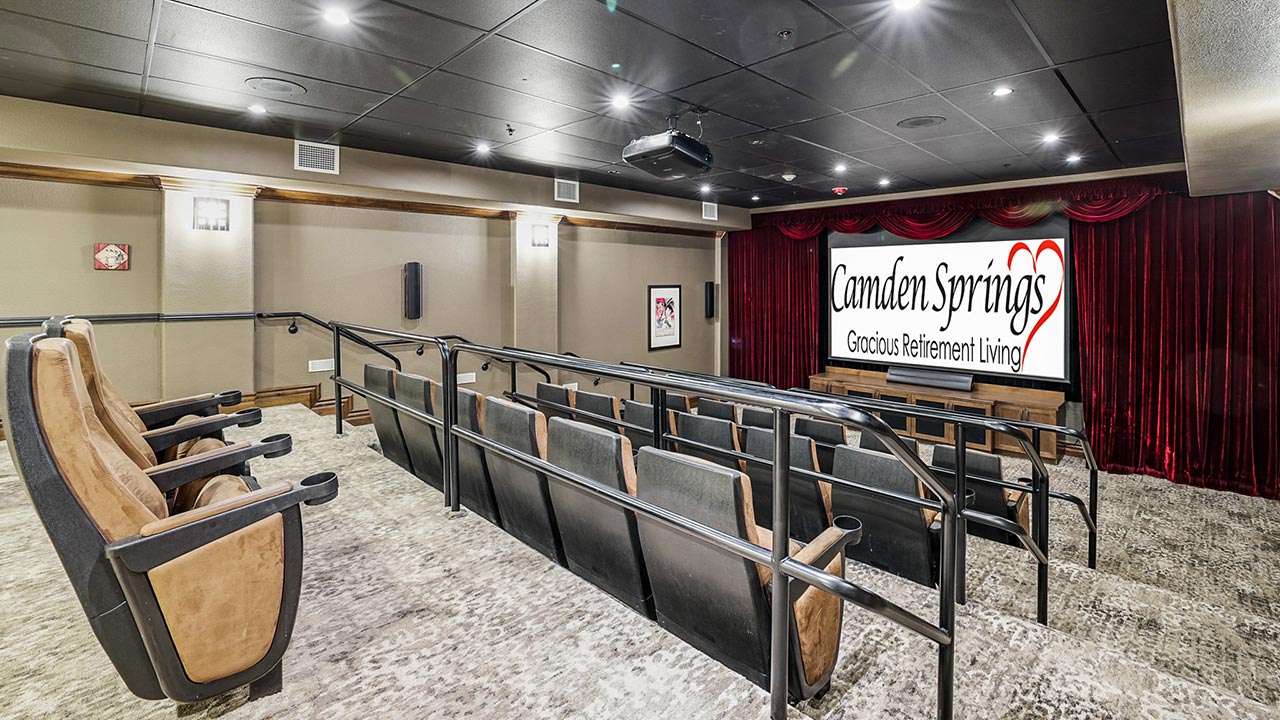 A small movie theater with tiered seating, brown cushioned chairs, gray carpeting, and a large screen displaying "Camden Springs Gracious Retirement Living" in front of red curtains.