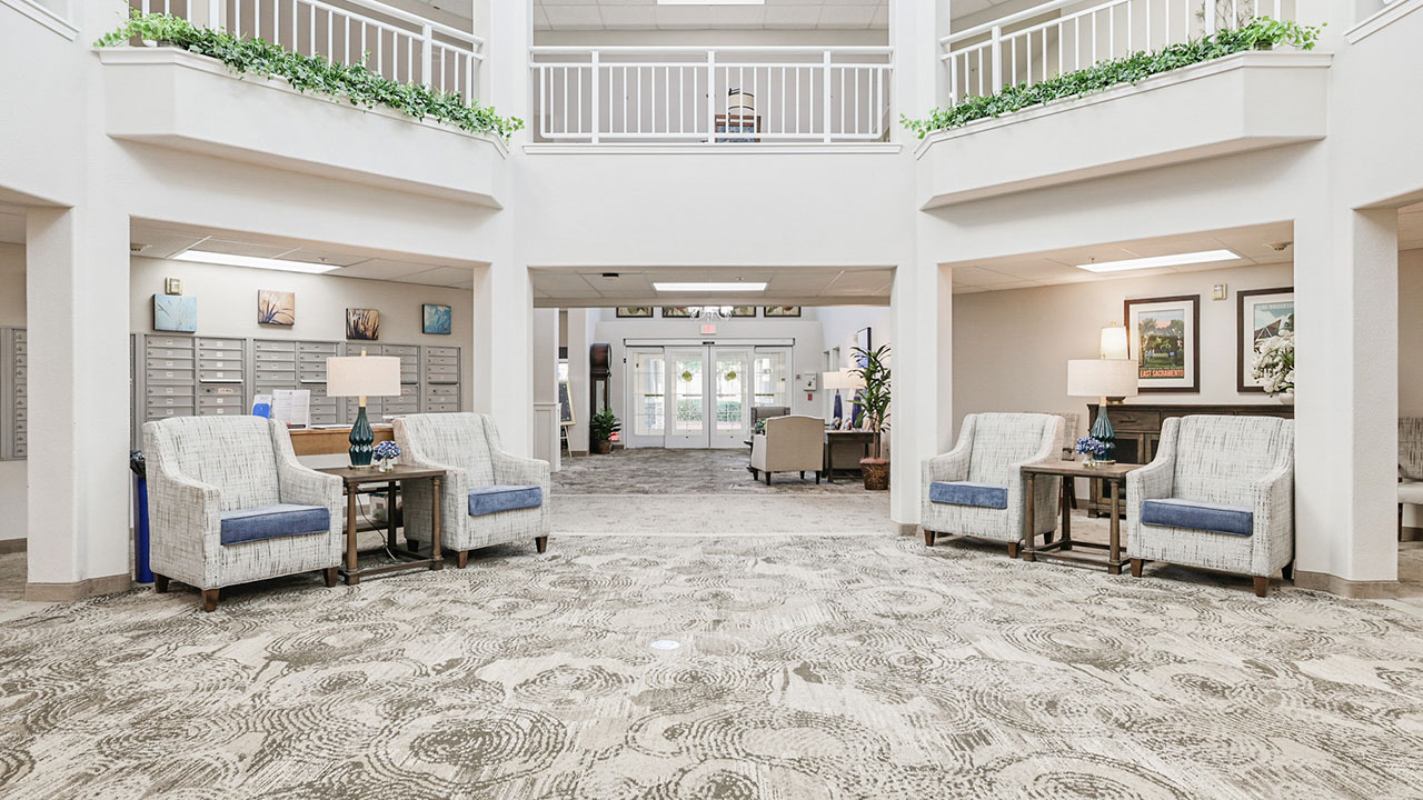 A bright, spacious lobby area with patterned carpet, two pairs of armchairs and tables on each side, indoor plants on the railings above, and mailboxes on the left wall. Large windows and glass doors let in natural light.