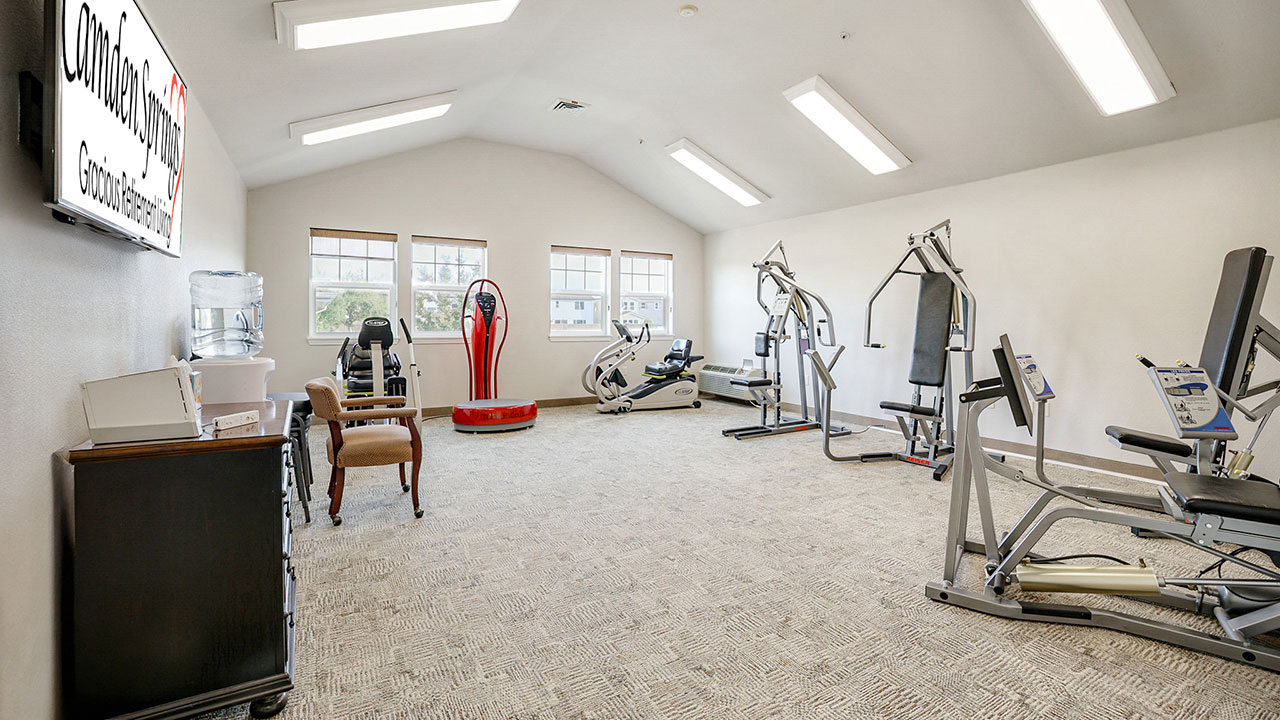 A bright, spacious gym room with various exercise machines, including treadmills, weight equipment, and recumbent bikes. There are chairs, a water dispenser, and large windows letting in natural light.