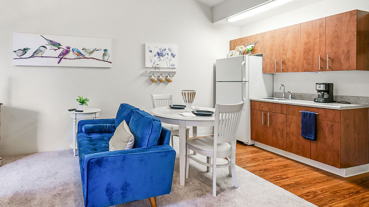A small open-concept kitchen and dining area with wood cabinets, white appliances, a round dining table with white chairs, and a blue velvet couch. Bird-themed artwork hangs on the walls.