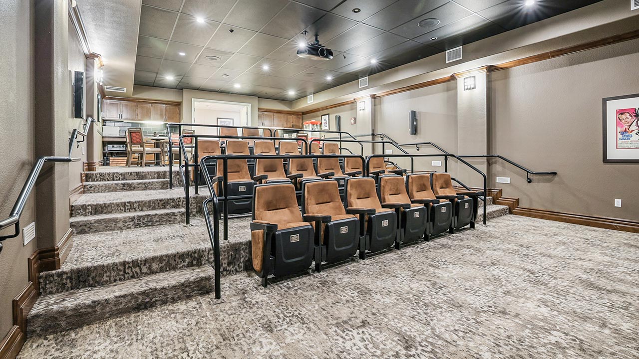 A modern home theater with three rows of brown reclining seats, carpeted floors, beige walls, and framed art. Black railings divide the seating, and a projector is mounted on the ceiling. A bar area is visible in the back.