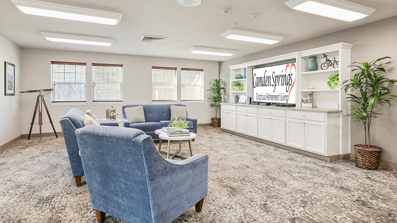 A spacious, well-lit living room features blue armchairs, a gray sofa, a round coffee table, and built-in white shelves. A large "Camden Springs Assisted Living" sign is displayed on the back wall.