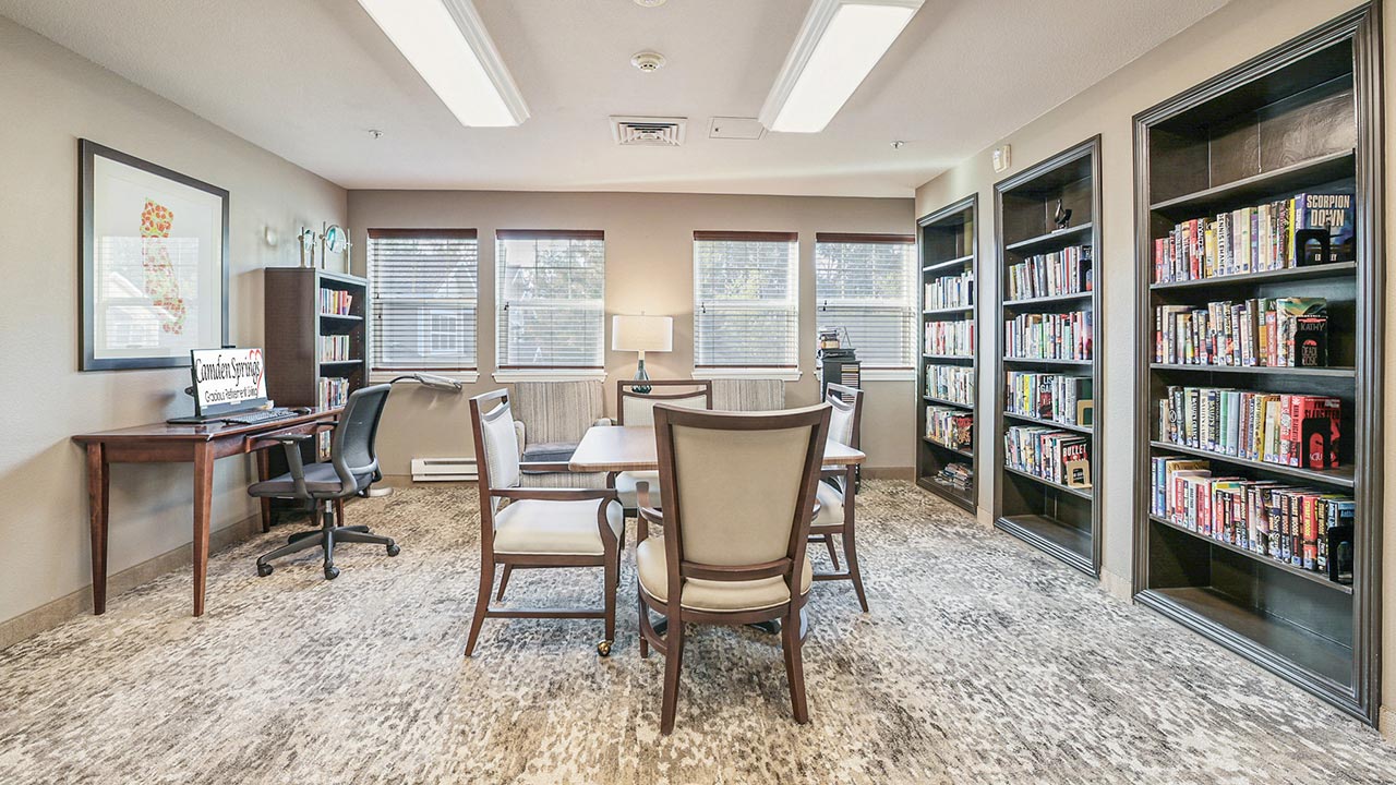 A bright room with a desk and computer on the left, a central table with chairs, bookshelves filled with books on the right, and large windows letting in natural light.