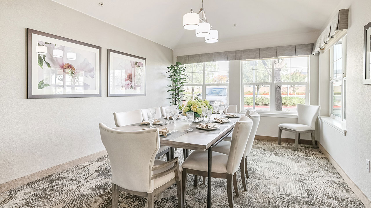 A bright, elegant dining room with a rectangular table set for six, cream-colored chairs, large windows letting in natural light, floral artwork on the wall, and a patterned beige carpet.