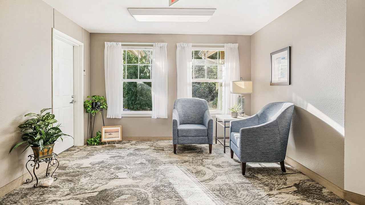 A bright, cozy room with two blue armchairs around a small table, a lamp, decorative plant, framed photo on the floor, and two large windows letting in natural light. Light patterned carpet and beige walls.