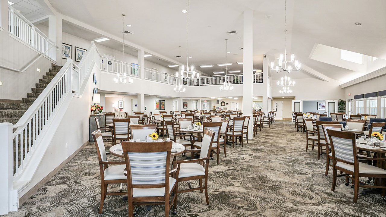 Spacious, well-lit dining room with round tables and wooden chairs, decorated with small flower centerpieces. Chandeliers hang from the ceiling, and there is a staircase and second-floor balcony in the background.