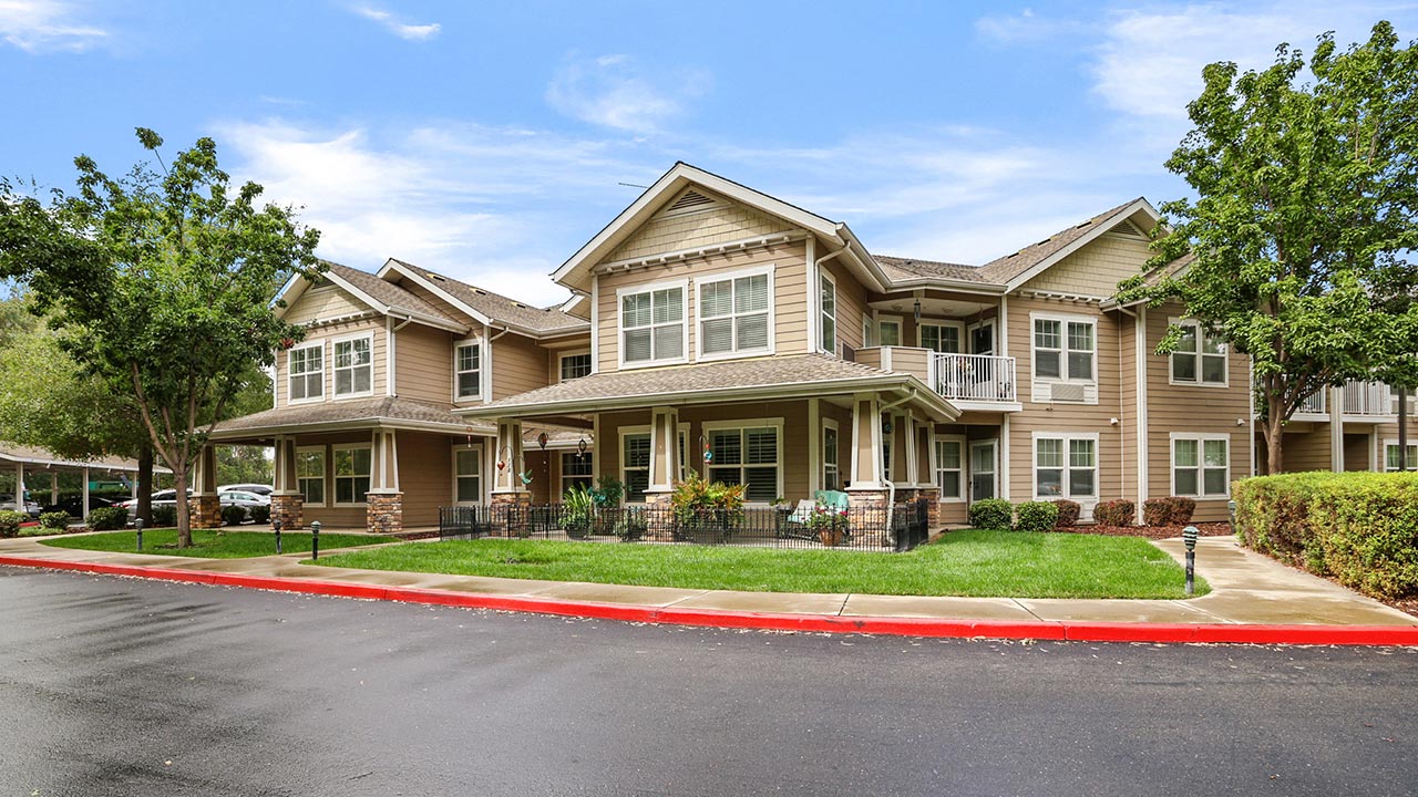 A large, two-story beige house with white trim, a manicured lawn, and trees, sits on a wet street with a red curb under a partly cloudy sky.