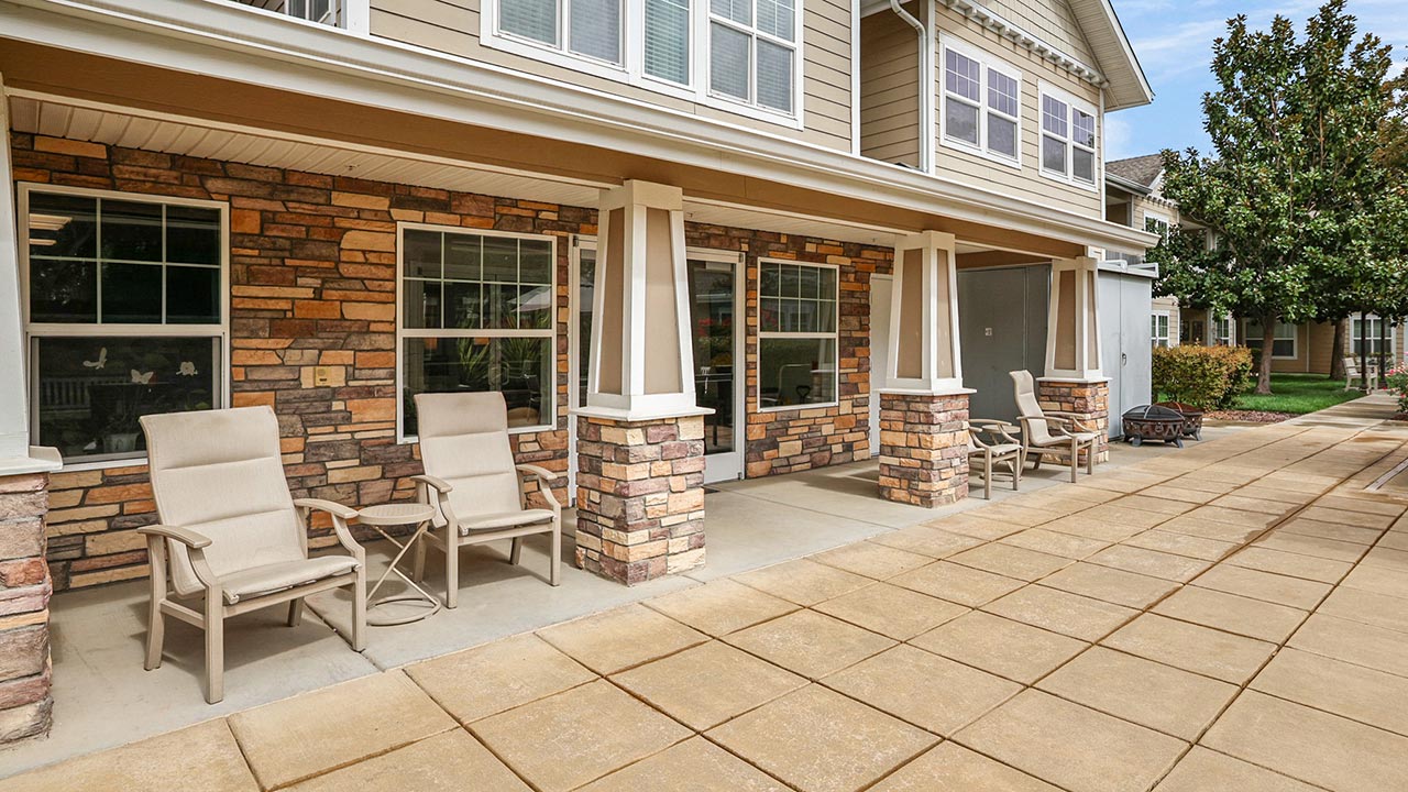 A spacious patio with beige lounge chairs and small tables sits in front of a building with stone and siding exterior, large windows, and white trim, next to a paved walkway and landscaped greenery.