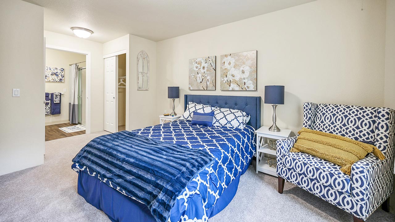 A bright bedroom with a blue and white bed, matching armchair, two nightstands with lamps, floral wall art above the bed, and a doorway leading to a bathroom in the background.