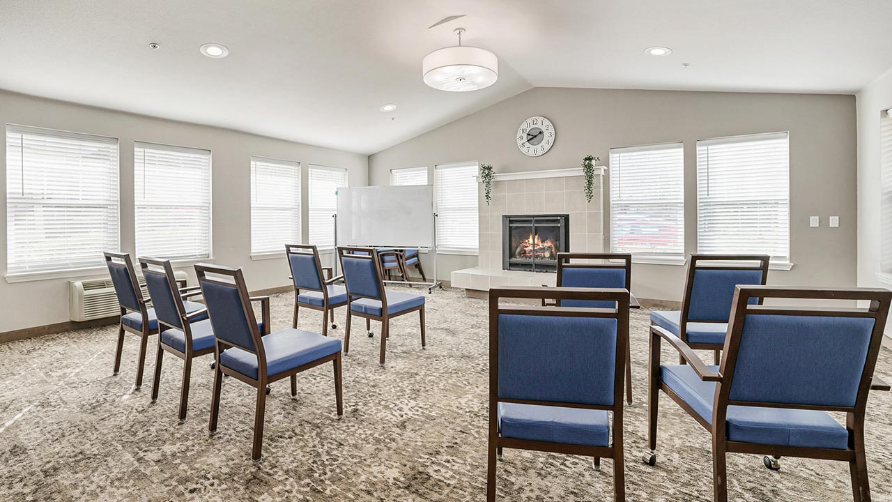 A bright room with several blue chairs arranged in a semi-circle facing a fireplace, whiteboard, and clock on the wall. Natural light streams in through multiple windows, creating a welcoming atmosphere.
