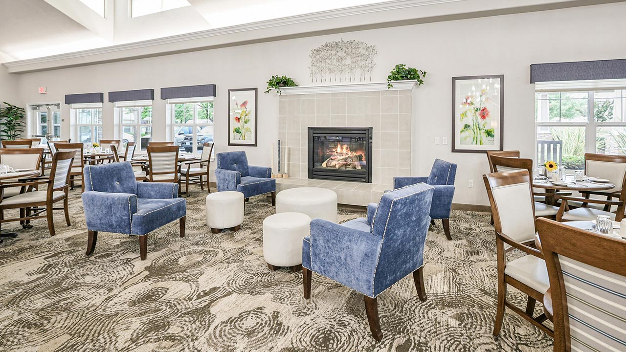 A bright, airy dining and lounge area features blue upholstered chairs, round white ottomans, wooden dining tables, a fireplace, large windows, and floral decor on the walls.
