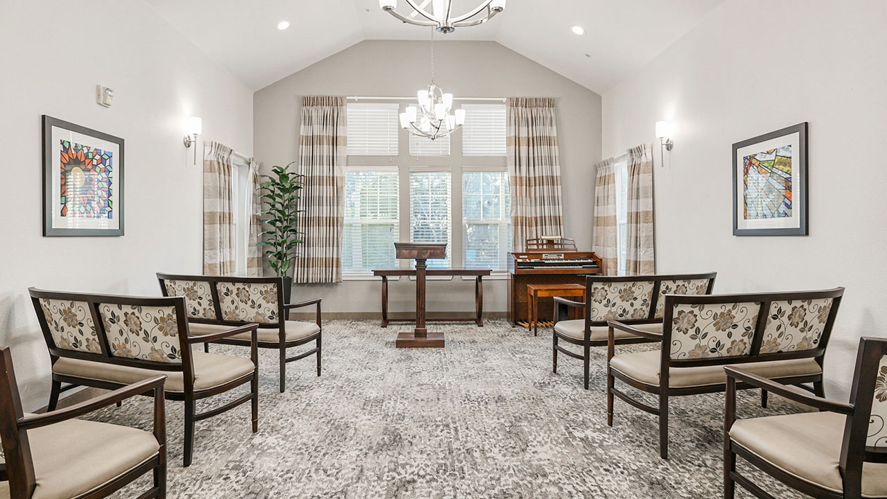 A small, bright chapel with patterned chairs in rows, a wooden podium, organ, and modern chandelier. Large windows with striped curtains let in natural light. Two framed artworks hang on white walls.