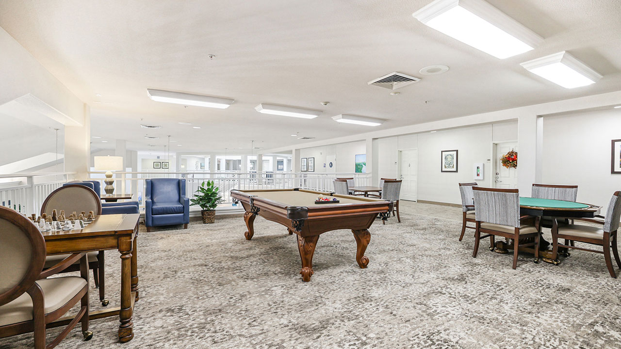 Spacious, well-lit game room with a pool table in the center, chess and card tables around, cushioned chairs, and framed art on white walls. Large windows provide natural light.