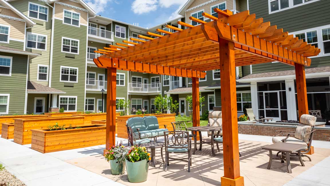 A courtyard with a wooden pergola featuring patio furniture underneath. Raised garden beds are lined up near the pergola, and a multi-story residential building with balconies is in the background. Flower pots and greenery add to the outdoor setting, providing an ideal independent living space.