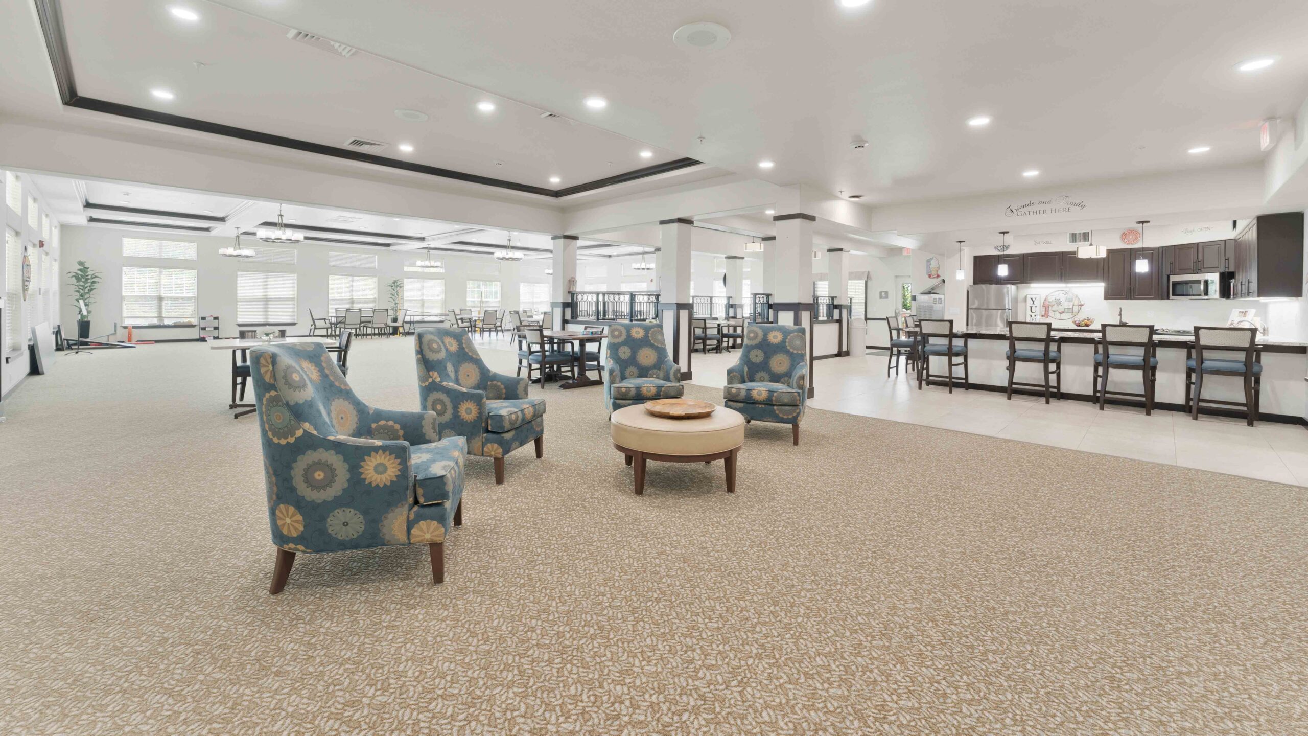 Spacious, well-lit lounge area with patterned carpet, three blue armchairs around a round coffee table, and dining tables in the background near a kitchen or café.