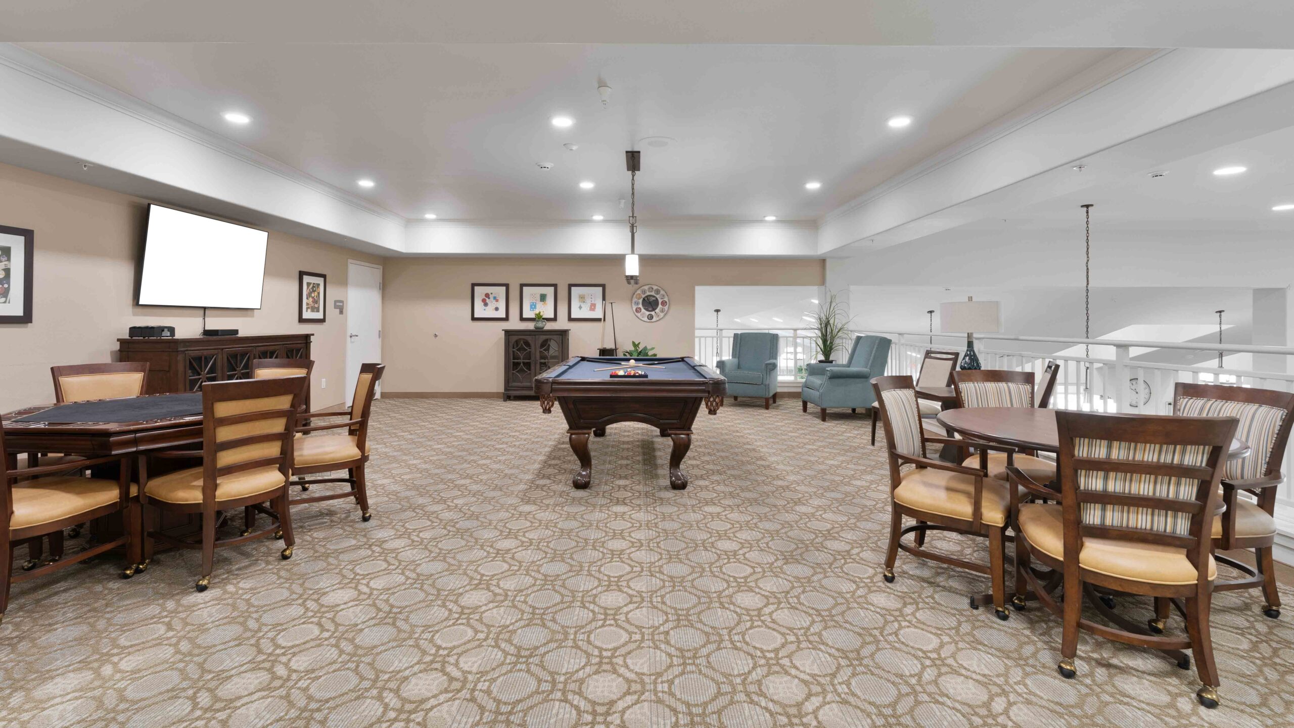 Spacious game room with a pool table in the center, card tables with chairs, lounge chairs near the wall, and framed art on beige walls. The room is well-lit with recessed lighting.