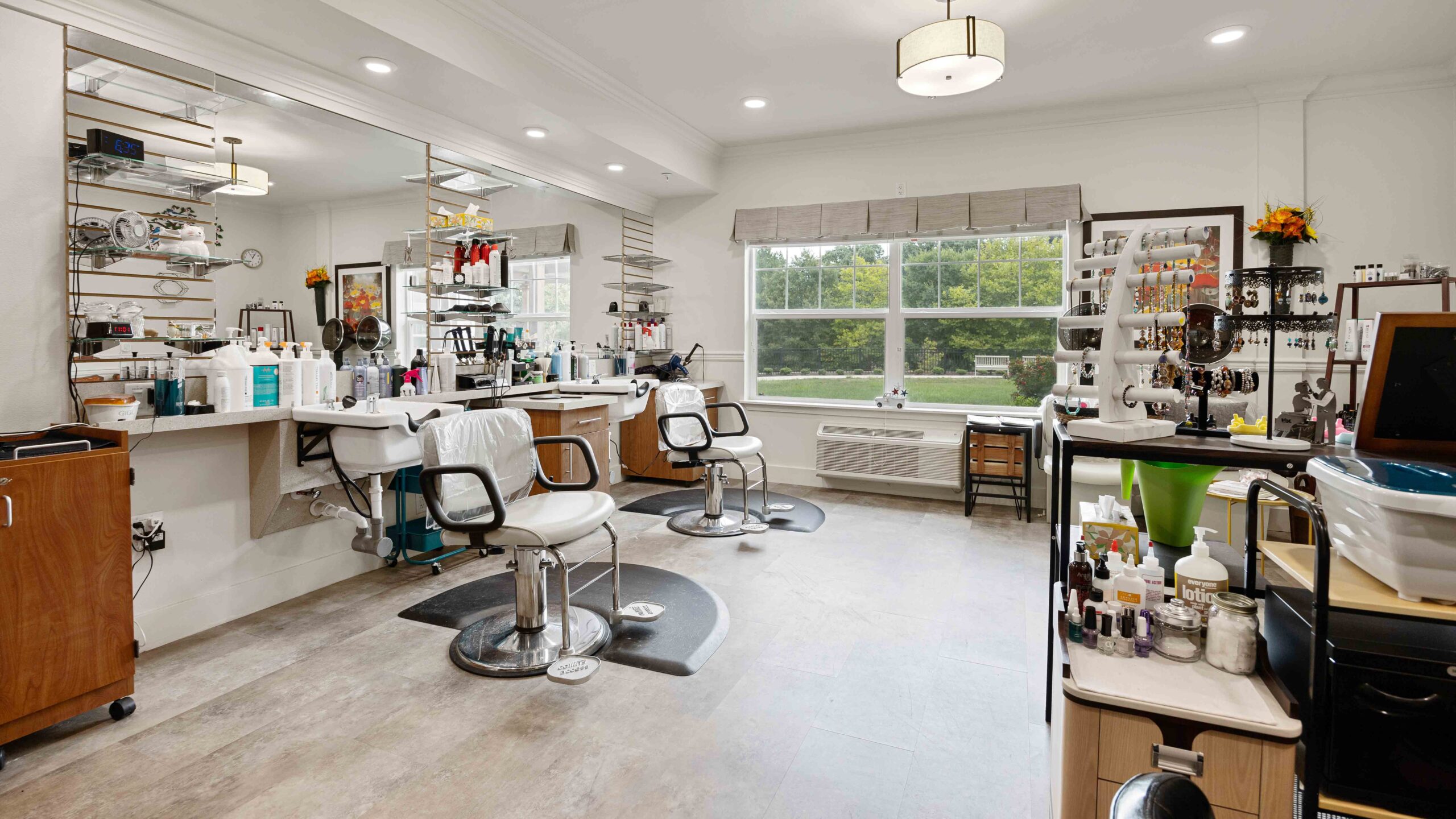 Bright, modern salon with two styling chairs in front of mirrors, shelves of hair products and tools, and a large window letting in natural light. The space is tidy and well-organized with various salon equipment.