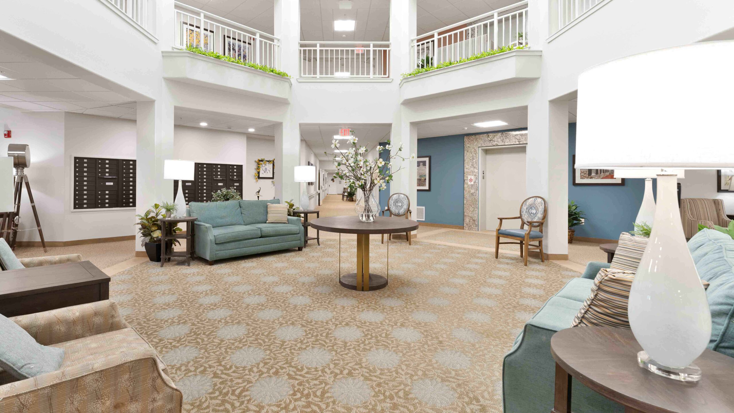 Spacious, well-lit lobby with patterned carpet, light blue sofas, round table with flower arrangement, modern lamps, chairs, wall art, and mailboxes on the far left wall. White railings line the upper level.