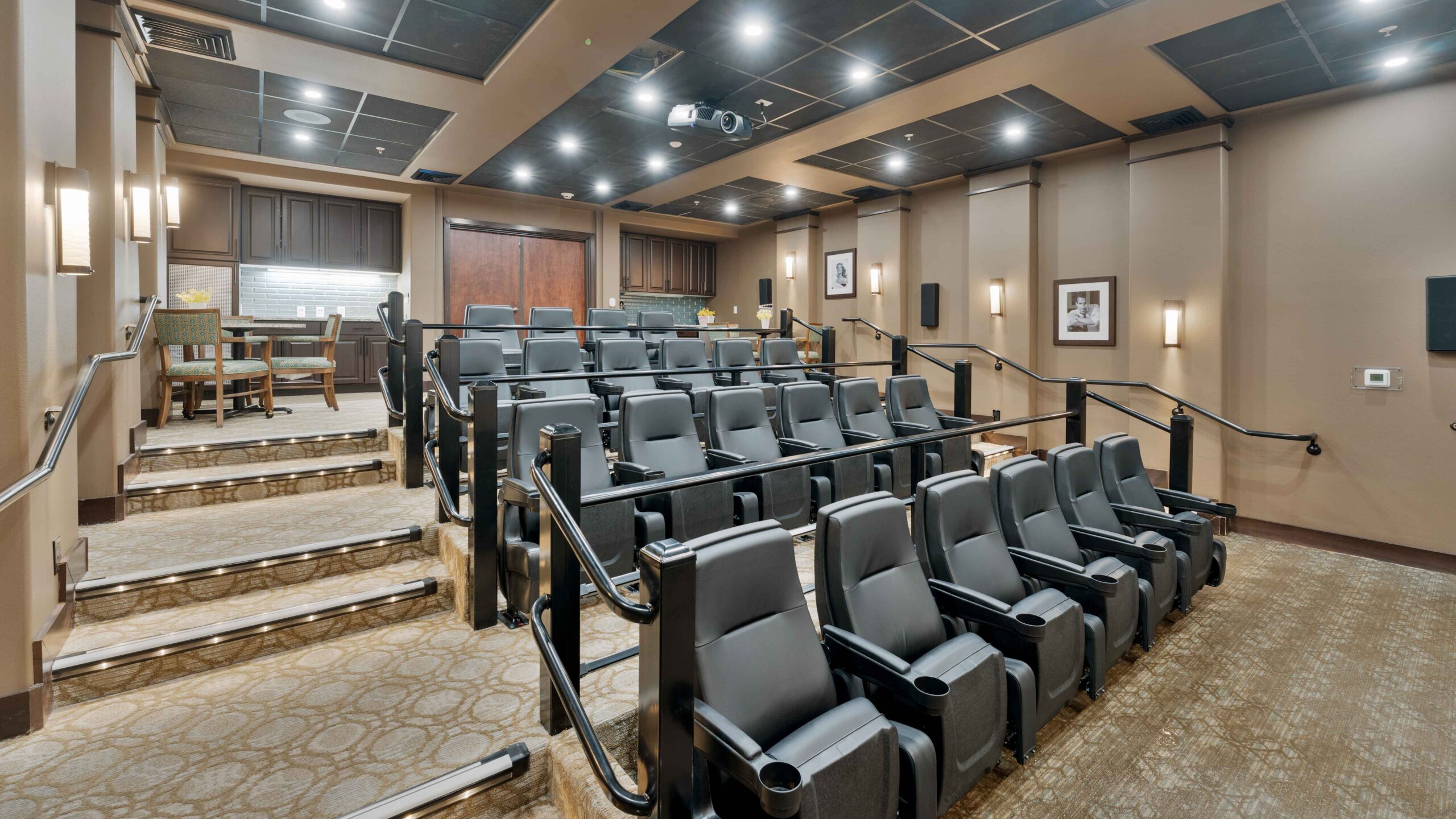 A small, modern theater room with several rows of black reclining seats, wall-mounted lights, carpeted floors, and a projector on the ceiling; a table and chairs are at the back of the room.