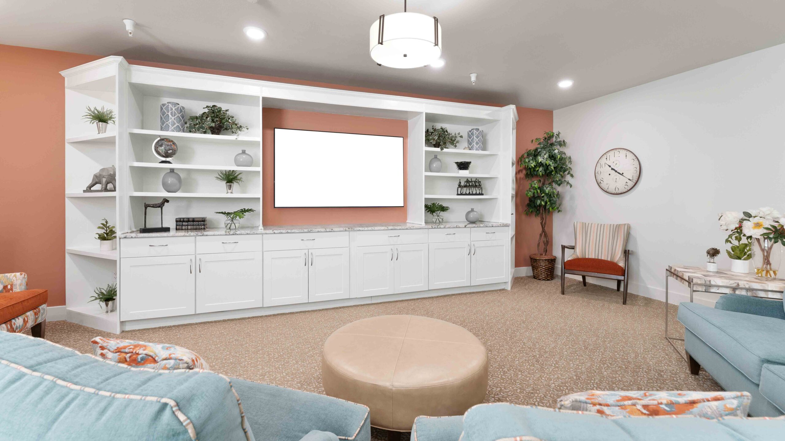 A modern living room with pastel blue sofas, a round beige ottoman, white built-in shelves with decor, a blank TV screen, an armchair, potted plants, a wall clock, and soft lighting.