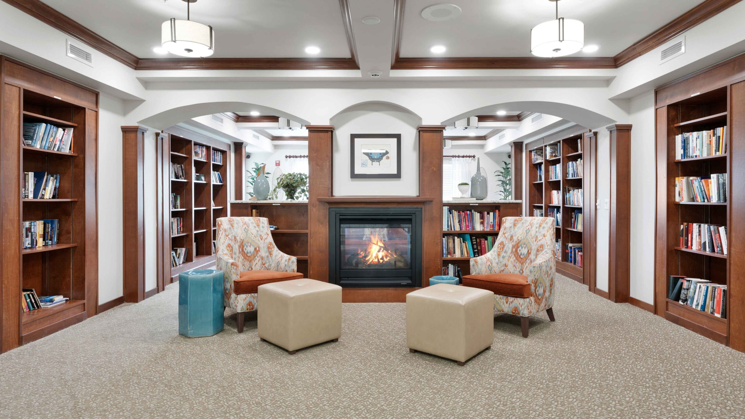 A cozy library with wooden bookshelves, two patterned armchairs, two beige ottomans, and a fireplace in the center. Modern light fixtures hang from the ceiling, and books line the shelves on all sides.