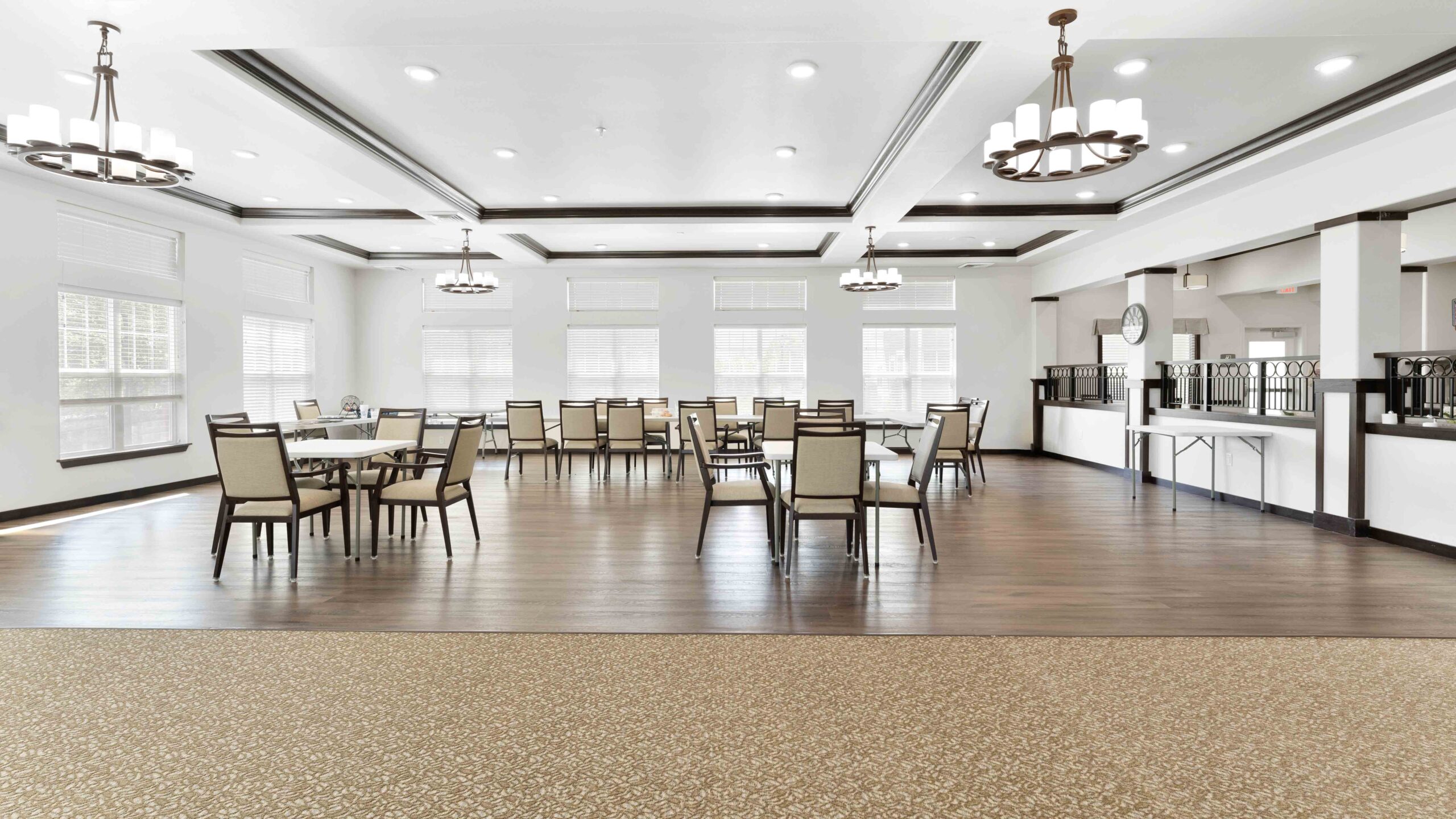 A spacious, well-lit dining room with wooden floors, several tables and chairs, large windows, and modern chandeliers hanging from the ceiling. The room has a clean, contemporary design.