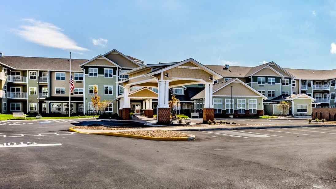 A modern, three-story assisted living facility with beige siding and white trim, featuring a covered entrance, large windows, landscaped grounds, and an adjacent empty parking lot under a clear blue sky.