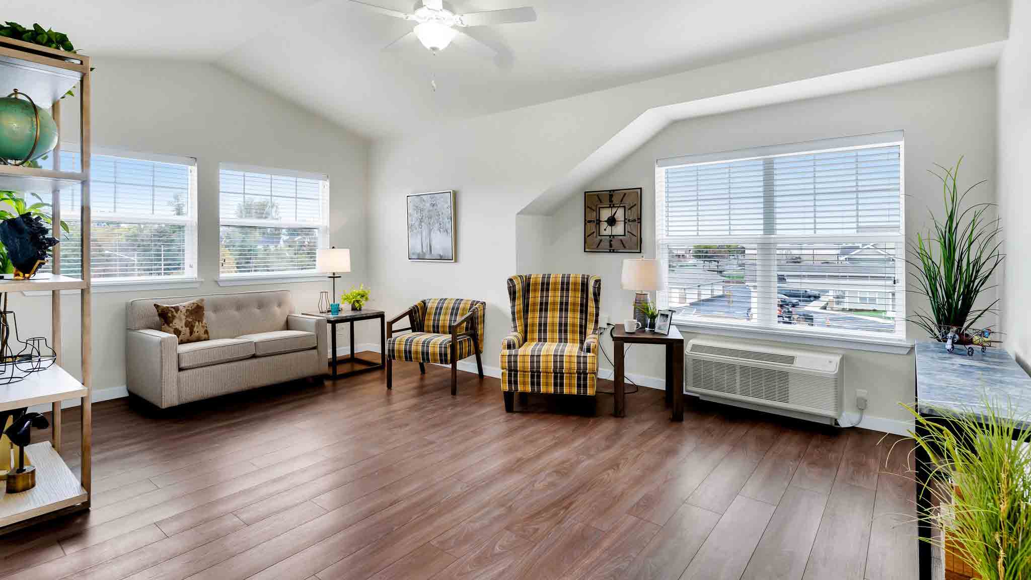 Bright living room with wood floors, beige sofa, two yellow plaid armchairs, large windows with blinds, plants, wall art, and a ceiling fan, creating a cozy and inviting atmosphere.