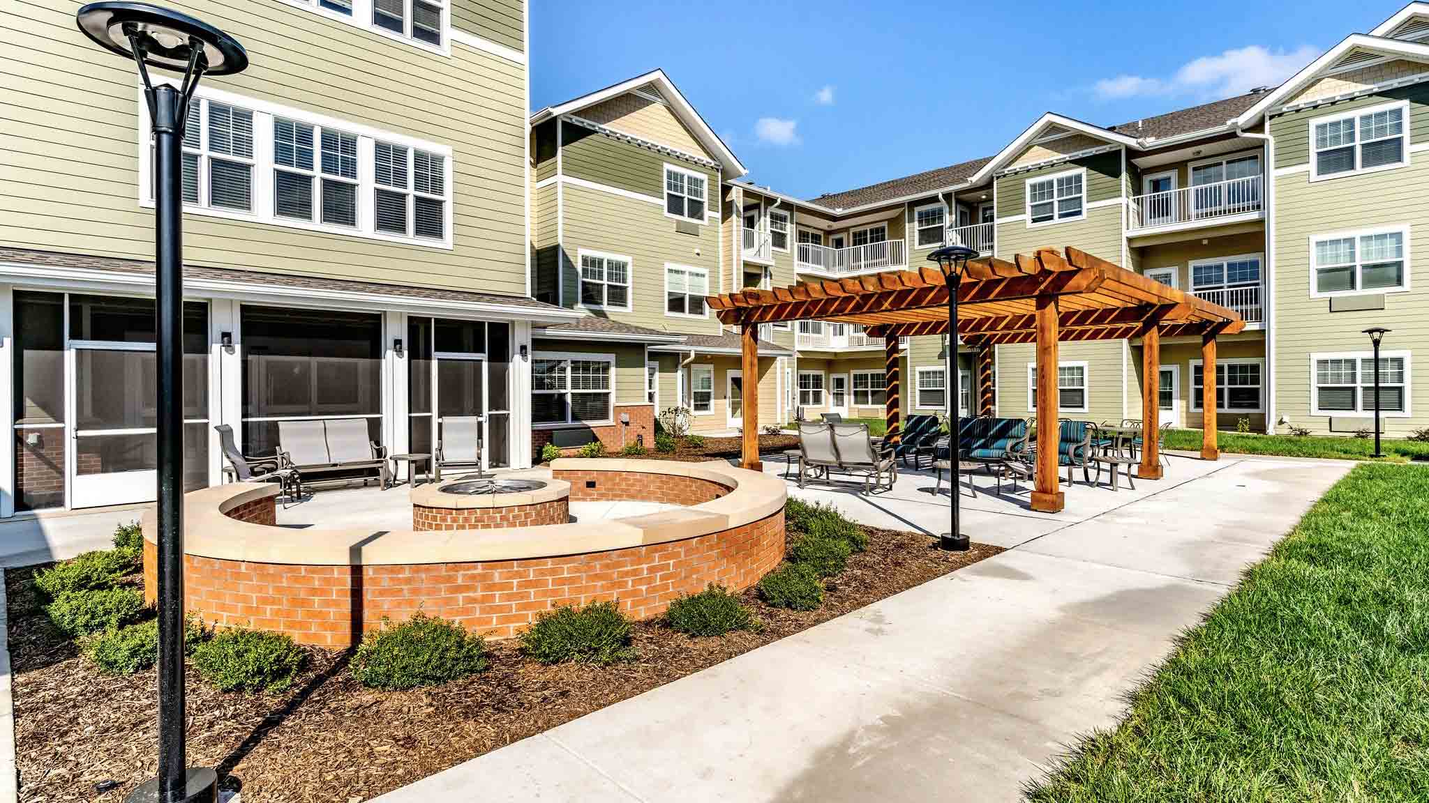 A modern apartment complex courtyard featuring a circular brick fire pit with seating, a wooden pergola with tables and chairs, sidewalks, green grass, shrubs, and multi-story beige buildings in the background.
