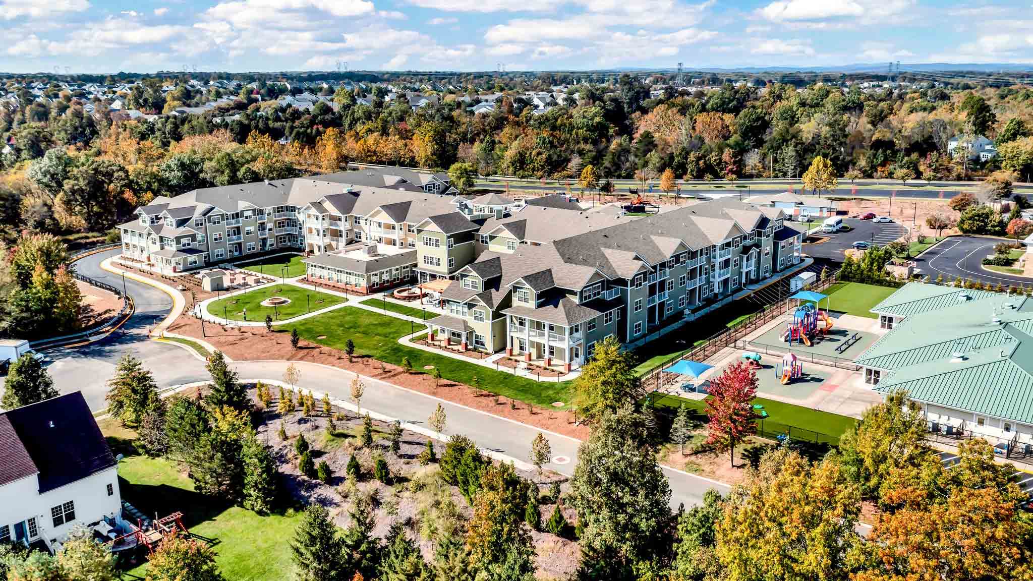 Aerial view of a large apartment complex with landscaped lawns, walking paths, and a playground, surrounded by trees with autumn foliage and nearby residential areas under a partly cloudy sky.