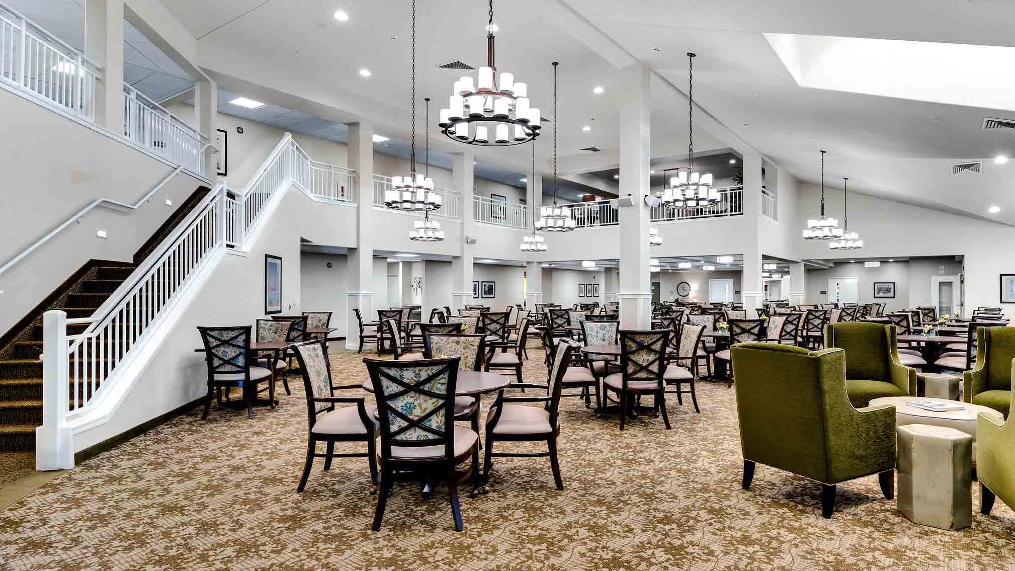 Spacious dining area with round tables and chairs, patterned carpet, armchairs on the right, white pillars, modern chandeliers, and a staircase leading to an upper level with railing. The room is brightly lit.